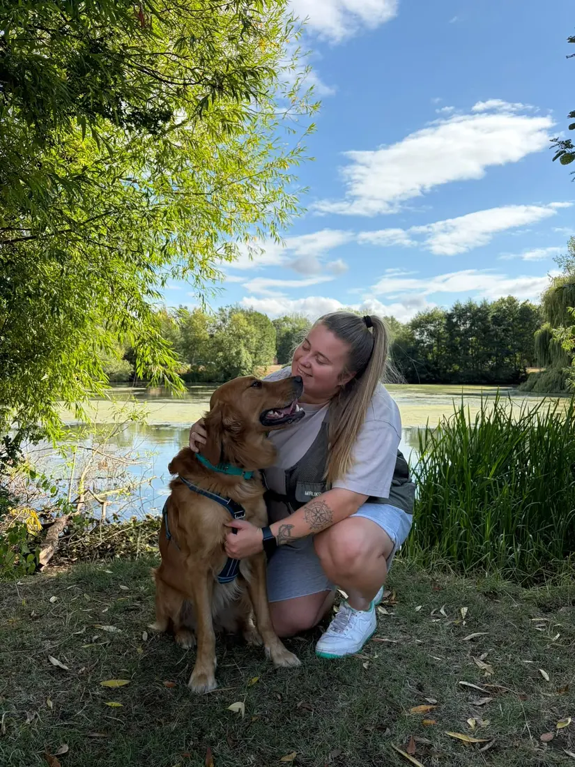 Aimee crouching by a lake with Oakley, her working-line Golden Retriever, who is looking up at her